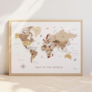 May include: A framed world map with a light wood frame. The map features a neutral color palette with shades of brown and beige, and the text "MAP OF THE WORLD" is displayed in the center. A compass rose is in the lower left corner.