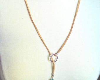 May include: A tan suede cord necklace featuring a silver-toned circular ring connector and a single teardrop-shaped blue stone pendant. The cord wraps around the neck and threads through the metal ring, with a small silver bead accenting the stone.