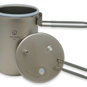 Puede incluir: Una olla de camping de titanio con tapa y asas plegables. La olla es cil&iacute;ndrica con un borde de silicona gris y la tapa tiene un asa. La olla tiene la marca "VALTCAN" impresa en el lateral.
