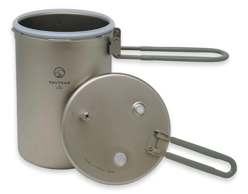 Puede incluir: Una olla de camping de titanio con tapa y asas plegables. La olla es cil&iacute;ndrica con un borde de silicona gris y la tapa tiene un asa. La olla tiene la marca "VALTCAN" impresa en el lateral.