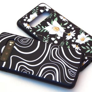 May include: Two black phone cases. One features a white, flowing, abstract design. The other has a floral pattern with white flowers and green leaves. Both cases have a camera cutout and are on a white background.