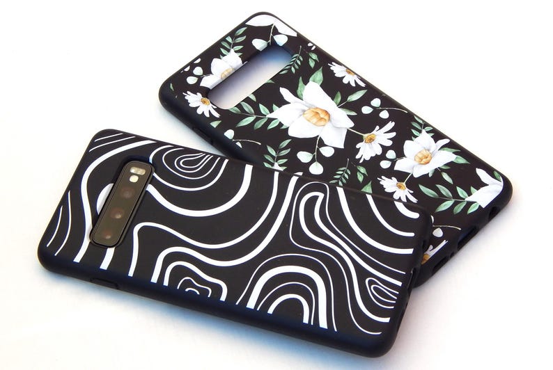 May include: Two black phone cases. One features a white, flowing, abstract design. The other has a floral pattern with white flowers and green leaves. Both cases have a camera cutout and are on a white background.