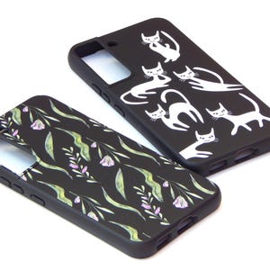 May include: Two black phone cases. One features white cat illustrations in various poses. The other has a floral pattern with green leaves and purple flowers. Both cases are on a white background.