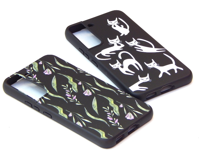 May include: Two black phone cases. One features white cat illustrations in various poses. The other has a floral pattern with green leaves and purple flowers. Both cases are on a white background.
