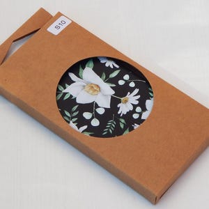 May include: A brown cardboard box with a circular cutout revealing a floral pattern. The pattern features white flowers and green leaves on a black background. A small white sticker with the text "S10" is on the box.
