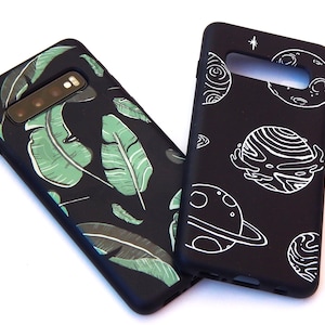 May include: Two black phone cases. One features a green and white leaf design. The other has a white planet and star design. Both cases are shown on a white background.