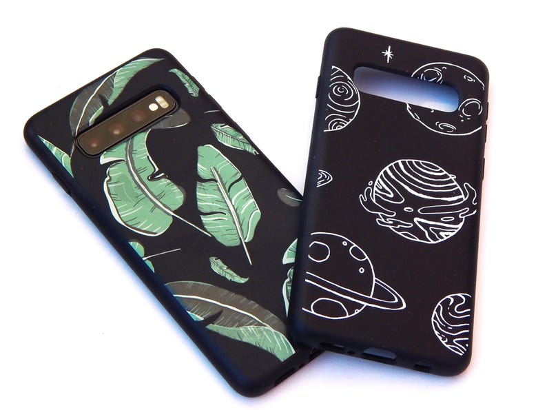 May include: Two black phone cases. One features a green and white leaf design. The other has a white planet and star design. Both cases are shown on a white background.