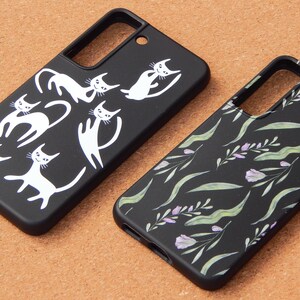 May include: Two black phone cases. One features white cat illustrations in various poses. The other has a floral pattern with green leaves and purple flowers. Both cases are on a brown surface.
