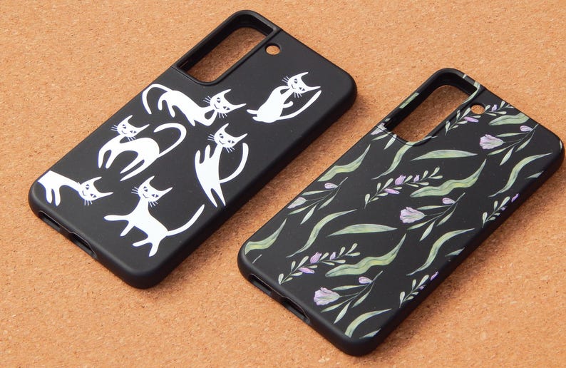 May include: Two black phone cases. One features white cat illustrations in various poses. The other has a floral pattern with green leaves and purple flowers. Both cases are on a brown surface.