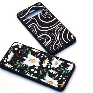 May include: Two black phone cases. One features a white, swirling pattern. The other has a floral design with white flowers, green leaves, and yellow centers on a black background. Both cases have camera cutouts.