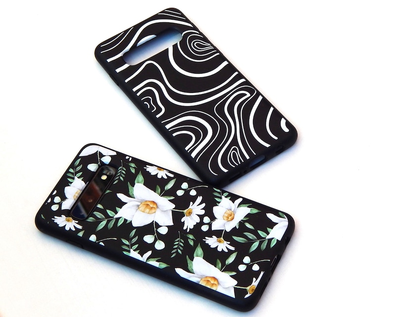 May include: Two black phone cases. One features a white, swirling pattern. The other has a floral design with white flowers, green leaves, and yellow centers on a black background. Both cases have camera cutouts.