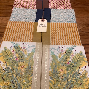 May include: A collection of five hardcover books arranged in a row, featuring diverse patterned covers including floral, geometric, and botanical prints. A small white tag with the handwritten text #1 is placed on the fourth book from the front.