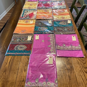 May include: A table display of twenty-one colorful saris, each wrapped in plastic and labeled with a number. The saris are arranged in rows, with the numbers increasing from left to right. The saris are a variety of colors, including pink, orange, yellow, green, and blue. Some of the saris have intricate embroidery and beading.
