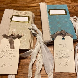 May include: Two handmade journals with fabric spines and metal label holders. The left journal has a beige patterned cover, and the right has a blue patterned cover. Each features a white fabric tie and a paper tag with handwritten text.