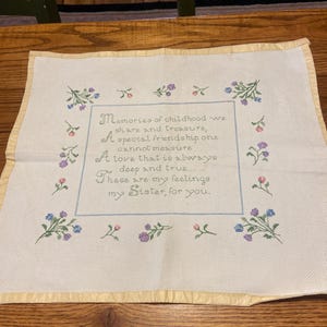 May include: A square, off-white embroidered piece with a light blue border and floral accents. The text reads, "Memories of childhood we share and treasure, A special friendship one cannot measure, A love that is always deep and true... These are my feelings my Sister, for you."