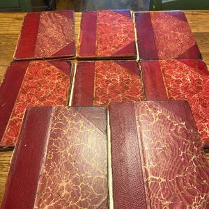 May include: A collection of seven antique books arranged in a grid. Each book features a deep red leather spine and corners, paired with a marbled paper cover in shades of red and gold. The books are rectangular with visible wear on the edges.