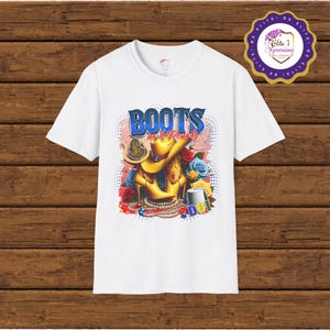 Peut inclure: T-shirt blanc avec un motif graphique coloré représentant une paire de bottes jaunes avec des perles et le texte "BOOTS and Pearls".
