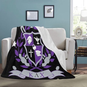 May include: A black and purple fleece blanket with a crest featuring a white eagle, a lamp, a padlock, a cross, and a stack of books. The crest is surrounded by a purple wreath and a banner with the letters "ΚΛΧ".