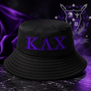 May include: A black bucket hat featuring the Greek letters KΛX embroidered in a vibrant purple color on the front. The hat has a flat crown and a downward-sloping brim with visible stitching detail around the circumference of the brim.