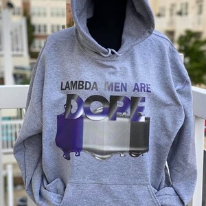 May include: A gray hooded sweatshirt with the text "LAMBDA MEN ARE DOPE" in a purple and silver design. The sweatshirt has a front pocket and a drawstring hood. The image is taken outdoors.