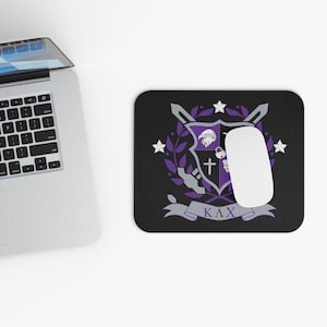 May include: Black rectangular mousepad featuring a purple and gray crest design with a cross, eagle, and stars. A white computer mouse rests on the pad. The Greek letters ΚΛΧ are at the bottom.