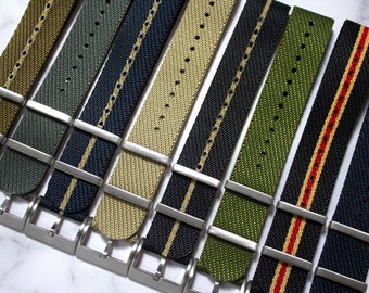 May include: A collection of eight nylon watch straps arranged in a row, featuring various colors including olive, navy, black, and tan, some with central stripes. Each strap includes a silver-toned metal buckle and two matching metal keepers.