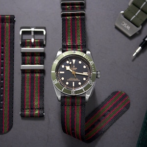 May include: A black and green striped watch strap with a silver buckle. The strap is made of nylon and has a red stripe running down the center. The strap is shown on a watch with a black face and green bezel.