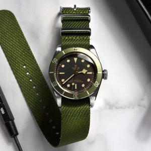 May include: A silver watch with a black face and green bezel on a green NATO strap. The watch face has white markings and the text "TUDOR" and "Oyster Prince" printed on it.