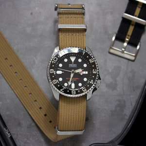 Peut inclure: Une montre-bracelet noire et argentée avec un cadran noir et des marquages blancs. La montre a un bracelet en nylon marron et se trouve sur une surface grise.