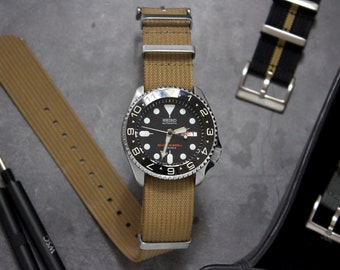 Könnte beinhalten: Eine schwarz-silberne Armbanduhr mit schwarzem Zifferblatt und weißen Markierungen. Die Uhr hat ein braunes Nylonarmband und liegt auf einer grauen Oberfläche.
