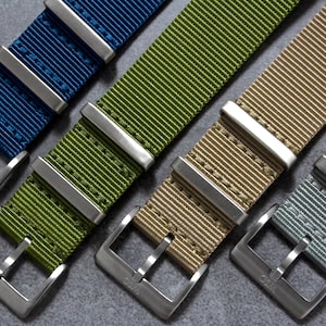 Pode incluir: Quatro pulseiras de relógio com fivelas prateadas. As pulseiras são de nylon e estão disponíveis em azul, verde, bege e cinza.