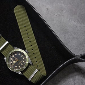 May include: A green nylon watch strap with a silver buckle, shown on a black watch with a green bezel and black face. The watch and strap are on a black leather watch tray.