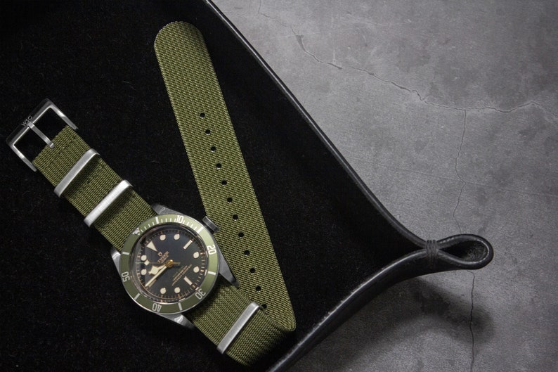 May include: A green nylon watch strap with a silver buckle, shown on a black watch with a green bezel and black face. The watch and strap are on a black leather watch tray.