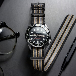 May include: A black wristwatch with a black face and silver hands. The watch has a black and tan striped nylon strap. The watch is on a gray surface with a pair of glasses and a tool.