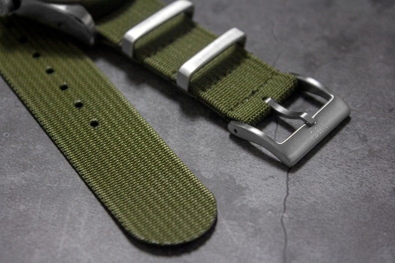 May include: A green nylon watch strap with a silver buckle. The strap has a textured surface and is designed for a watch with a 20mm lug width.