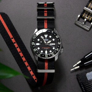 May include: A black and red striped nylon watch strap with a silver buckle. The strap is attached to a black watch with a silver bezel and a black face. The watch face has white markings and a date window.