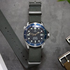Op de afbeelding: Een zilveren Omega Seamaster Professional 300m horloge met een blauwe bezel en een zwarte wijzerplaat. Het horloge is op een grijze NATO-band.