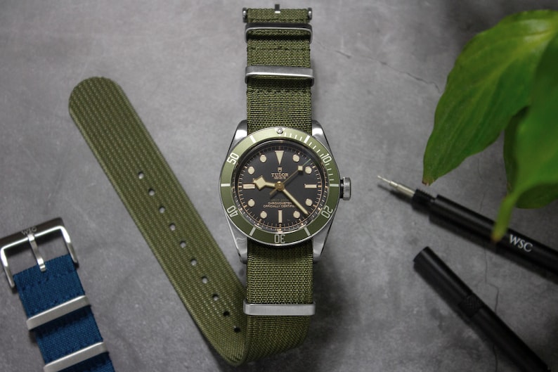 May include: A silver watch with a black face and green bezel on a green nylon strap. The watch has a date window at the 3 o'clock position and a seconds hand. The strap has a buckle closure.