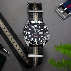 Könnte beinhalten: Eine Seiko Automatikuhr mit schwarzem Zifferblatt, leuchtenden Indizes und einer Tages-Datums-Anzeige. Die Uhr verfügt über eine drehbare Lünette mit weißen Ziffern und ein schwarz-grau-beigefarbenes Nylonarmband mit silberfarbenen Beschlägen.