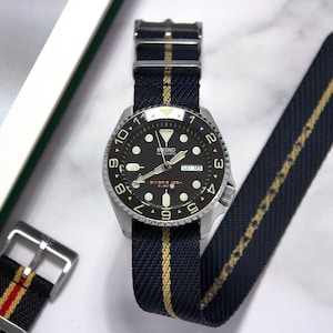 Puede incluir: Un reloj Seiko negro y plateado con una correa de nylon negra con una raya dorada. La esfera del reloj tiene un fondo negro con números y manecillas blancas. La correa del reloj tiene un cierre de hebilla.