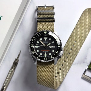 Puede incluir: Un reloj de buceo Seiko con esfera negra y correa de nailon color beige. El reloj tiene un bisel giratorio y marcadores luminosos. También se ven una correa de repuesto y una herramienta para relojes, junto con una caja Rolex.