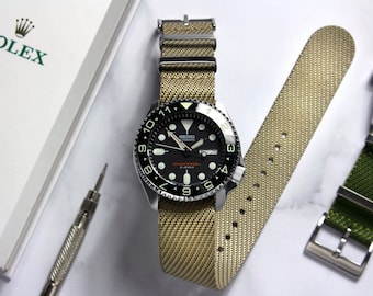 Op de afbeelding: Een Seiko duikershorloge met een zwarte wijzerplaat en een beige nylon band. Het horloge heeft een draaibare bezel en lichtgevende markeringen. Een reserveband en een horlogegereedschap zijn ook zichtbaar, samen met een Rolex-doos.