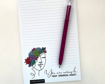 May include: A white lined notepad with a black and white line drawing of a person with a floral crown. The text "You are enough. Today. Tomorrow. Always." is printed below the drawing. A pink pen is lying on top of the notepad.