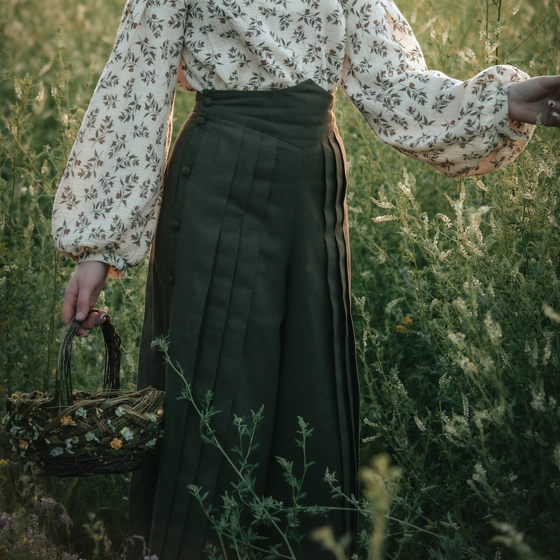 May include: A woman wearing a white blouse with a floral pattern and a dark green pleated skirt. She is holding a woven basket with flowers.
