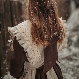 May include: A brown dress with a white lace overlay and a brown ribbon tied at the back. The dress is covered in snowflakes.