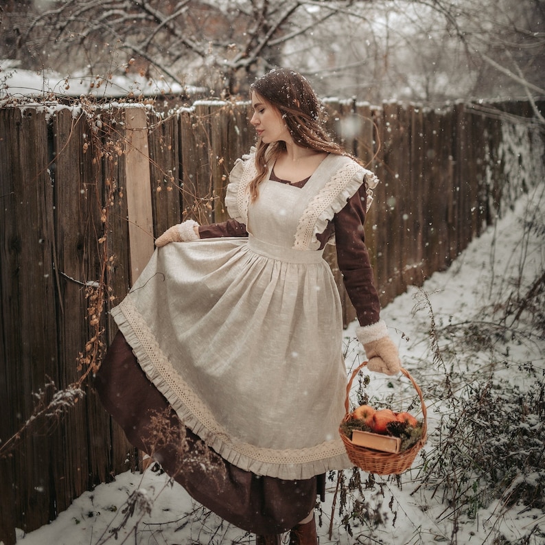 May include: A woman in a brown dress with a white apron and lace trim stands in a snowy yard. She is holding a basket of apples and a book. The background is a wooden fence and snow-covered trees.