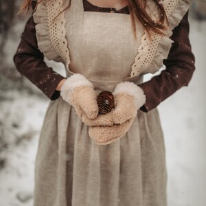 May include: A woman wearing a brown dress with a white lace overlay and white mittens holds a pine cone in her hands. The dress has a ruffled neckline and a gathered skirt. The woman is standing in a snowy forest.