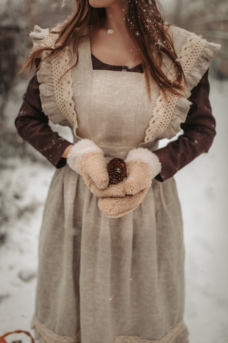 May include: A woman wearing a brown dress with a white lace overlay and white mittens holds a pine cone in her hands. The dress has a ruffled neckline and a gathered skirt. The woman is standing in a snowy forest.