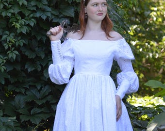 Op de afbeelding: Een witte off-the-shoulder jurk met lange, pofmouwen en een bloemenpatroon. De jurk wordt gedragen met een zilveren tiara en oorbellen. De achtergrond bestaat uit weelderig groen gebladerte.