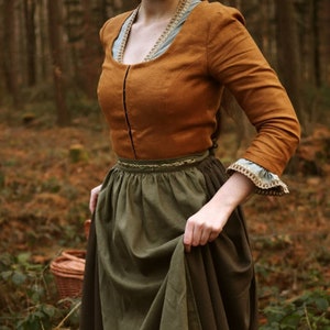 May include: A long-sleeved, rust-colored top with a scoop neckline and a dark vertical seam. The top is paired with a green skirt. The sleeves have a light blue lining and a decorative trim. The outfit is reminiscent of historical attire.